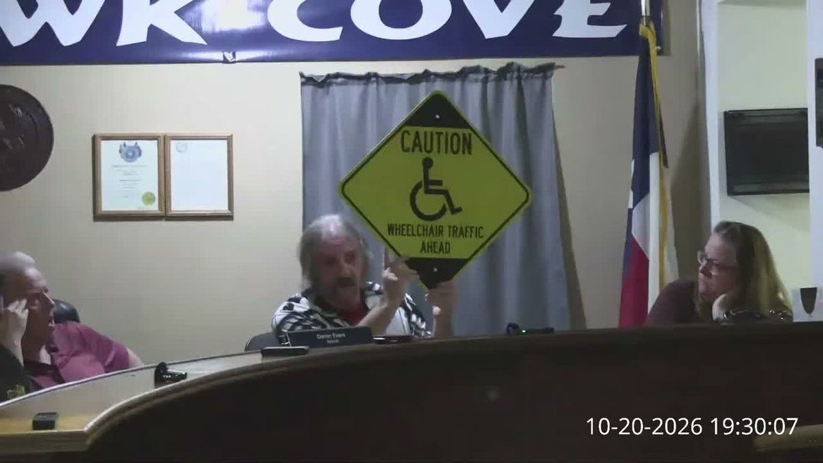 Council Member Kenneth Cork at the council dais holding up a yellow-green diamond sign reading CAUTION, with a wheelchair symbol and the words WHEELCHAIR TRAFFIC AHEAD